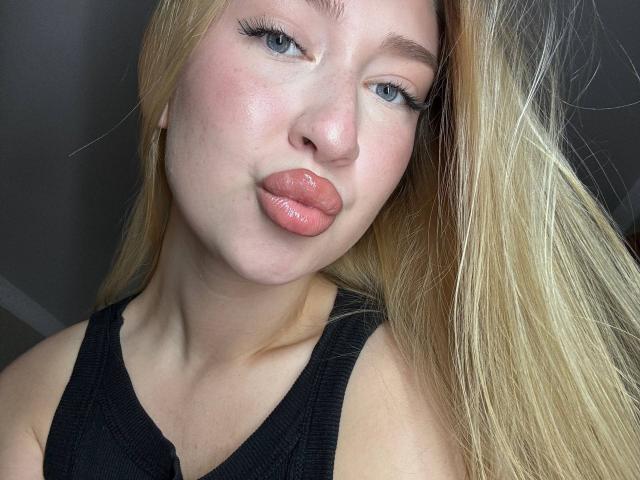 Joette Shigeta, met volle, glanzende, roze lippen, blond haar, blauwe ogen en een zwart tanktopje, kijkt in de camera met lichtjes open mond. Haar borsten zijn nauwelijks zichtbaar..