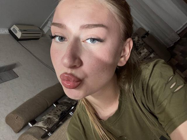Joette Shigeta, met volle, glanzende roze lippen, blauwe ogen en een blonde paardenstaart, draagt een groene blouse in een licht onscherpe selfie. Haar neus is klein en haar huid is licht..