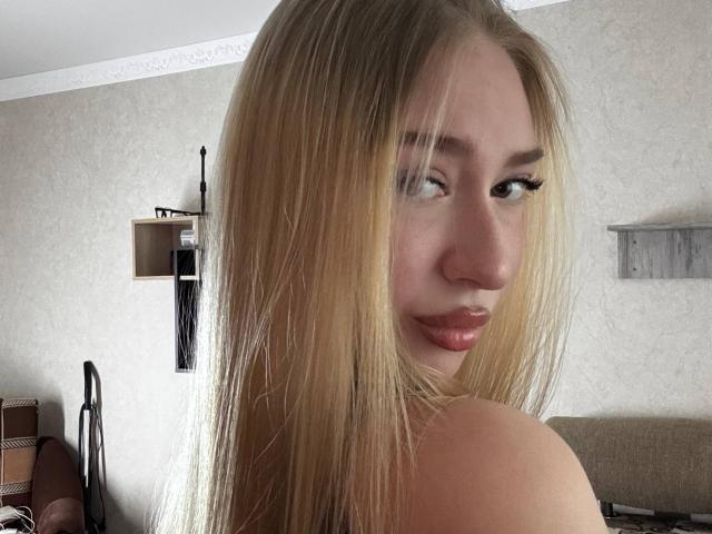 Joette Shigeta, blonde, avec de longs cheveux, de grosses lèvres et une forte poitrine, regarde par-dessus son épaule, montrant ses seins, dans une pièce confortable. Ses cils sont longs et ses yeux sont à moitié fermés..