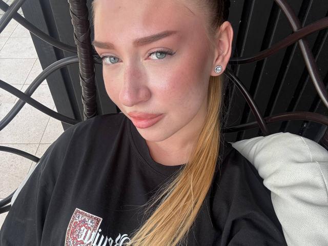 Joette Shigeta, met een blond staartje, draagt een zwarte top en heeft blauwe ogen, roze lippen en een diamanten oorbelle. Haar borsten zijn klein en haar gezicht is lichtelijk rood..