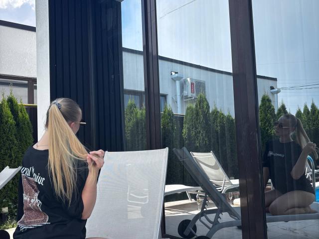 Joette Shigeta mit langem, blondem Pferdeschwanz, schwarzem T-Shirt, die einen weißen Netzstuhl justiert, Spiegelung im Glas, Außenbereich, sitzend auf einem weißen Stuhl, grüne Bäume, blauer Himmel, Sonnenbrille..