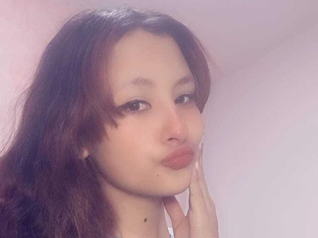 ValentinaRoseX with long brown hair, light skin, wearing a white graphic tee, showing her tits, and pink lips, touching her cheek with a hand, looking at the camera.