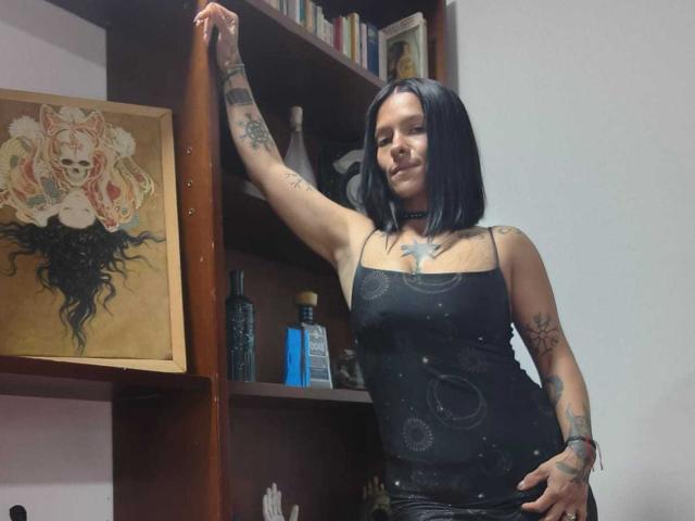 Amelie Blake, avec sa coupe de cheveux courte noire et ses tatouages, est appuyée contre une étagère en bois, vêtue d'un haut noir à bretelles fines, orné d'un motif galactique, qui révèle ses petites seins et un collier noir..