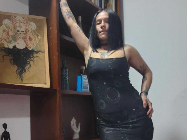 Amelie Blake, met haar zwarte haar en tatoeages, leunt tegen een houten plank, gekleed in een strakke zwarte jurk met hemelse patronen, met haar hand op haar heup, haar arm omhoog, en ze straalt zelfvertrouwen uit..