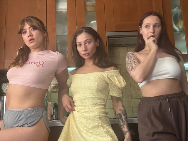 Three HotCaramelsTrio standing in a kitchen, wearing pink crop top and grey shorts, yellow off-shoulder dress, and white strap top with brown pants, showcasing different physiques.