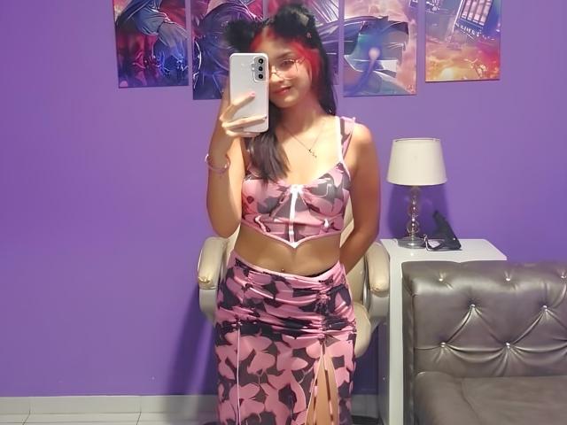 AriaRosel with pink and black floral outfit, showing her tits in a mirror selfie, wearing cat ears, glasses, and a smile, holding a phone in a purple room.