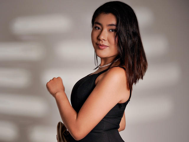 HannaGoez with dark hair, wearing a black dress, pearl necklace, and subtle makeup, standing with her right fist raised, showing her tits and a confident smile.