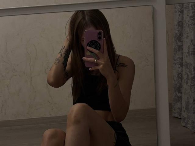 JaPorada, avec de longs cheveux bruns, un débardeur de sport noir et un short noir, assise, prenant un selfie devant un miroir avec un téléphone rose, tatouage sur le bras droit, jambes croisées..