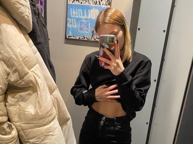 JaPorada mit blonden Haaren, einem schwarzen Crop-Top und einer schwarzen Hose mit hoher Taille, die in einem Spiegel-Selfie ihren nackten Bauch zeigt, hält ein Handy mit einer bunten Hülle..