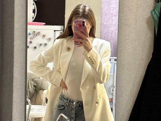 JaPorada, con lunghi capelli castani, indossa un cappotto giallo con bottoni dorati, una maglietta bianca e jeans blu, sta scattando un selfie allo specchio, mostrando una mano appoggiata al fianco e un telefono..