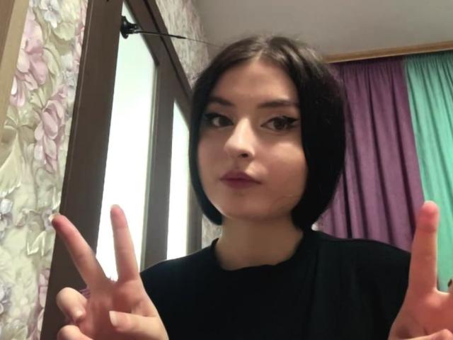 CindiMarkie met een zwart bob kapsel, draagt een zwarte blouse, maakt een dubbele v-teken met roze lippenstift, heeft een lichte huid, bruine ogen en subtiele make-up, en staat in een kamer met bloemen behang..