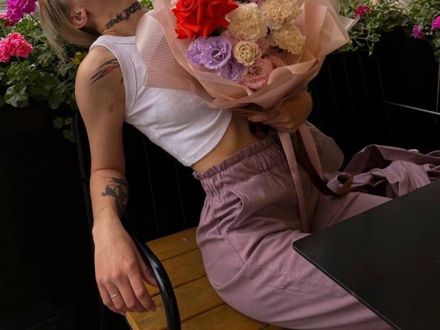 JaPorada mit einem Choker, einem weißen Crop-Top, rosa Hosen, die einen Blumenstrauß hält, Tätowierungen an den Armen, sitzt auf einer hölzernen Bank, rosa Blumen in der Hand..