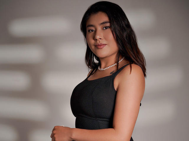 HannaGoez with medium tits, wearing a black tank top and pearl necklace, dark wavy hair, soft smile, looking at camera, standing against blurred background.