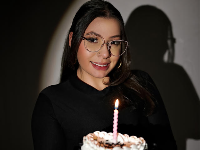 Vanessa, lunettes, souriante, haut noir, tenant un gâteau avec une bougie rose, ombre sur le mur, longs cheveux bruns, boucles d'oreilles cercles, piercing au nez, mignonne, jolie, fond sombre, éclairée par l'avant..