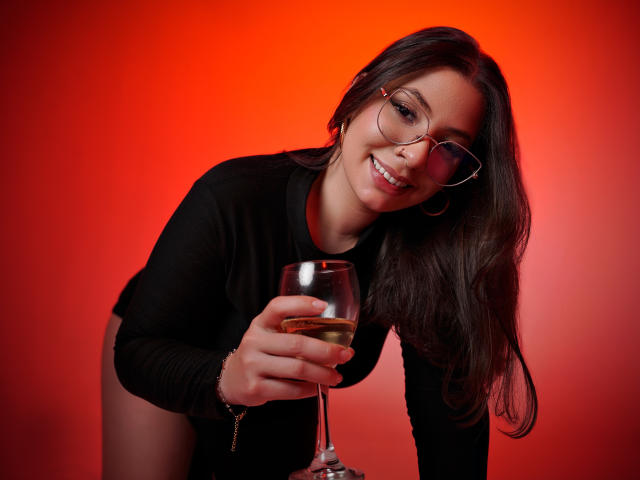VanessaClose with glasses, black long-sleeve top, holding a glass of white wine, smiling, leaning forward, red background, long brown hair, hoop earrings, visible butt.