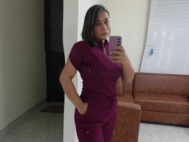 EmaFoxy with dark brown hair, wearing maroon scrubs, standing, hand in pocket, taking selfie in mirror, holding pink phone, brown leather couch in background.