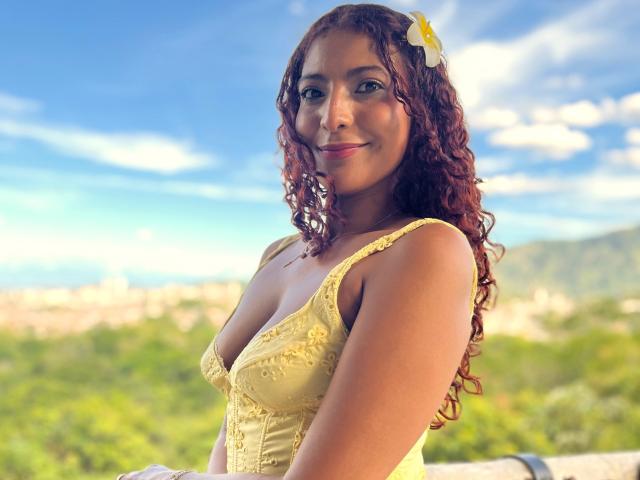 Mia Bianchi, con i capelli rossi ricci, un vestito giallo e un fiore bianco tra i capelli, sorride alla macchina fotografica, mostrando il seno, con un cielo azzurro brillante e un paesaggio verde sullo sfondo..