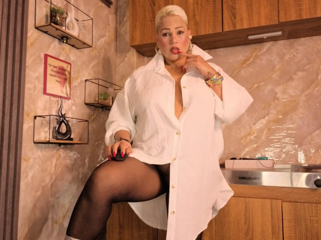Gianella Cox, con i capelli biondi, indossa una camicia bianca con bottoni, collant neri e rossetto rosso, ha una mano sulla bocca, con una gamba sollevata, mostrando la sua vulva..