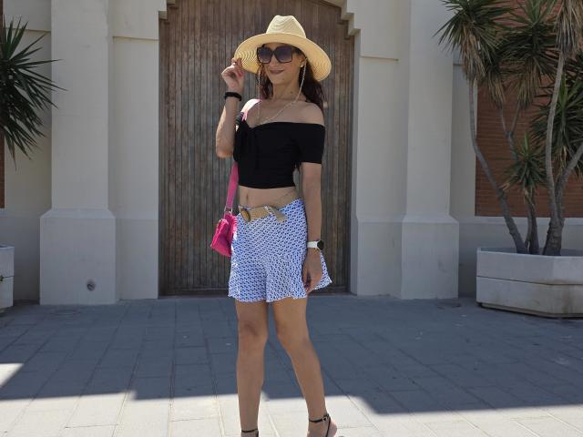 Cassandra Michelli draagt een zwart top met een open schouder, een blauw-witte rok, een bruine hoed, zonnebril, een roze tas, bruine benen en zwarte hakken, en staat met een houten deur achter zich..