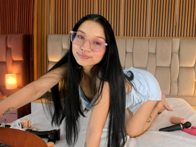SophieDelaney on all fours on a bed, wearing pink glasses, light blue shorts, long black hair, nose ring, tattoos on arm and leg, dildo and vibrator on bed.