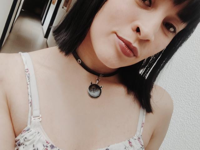 IssisDiosa a la peau pâle, les cheveux noirs mi-longs, et porte un collier noir avec un pendentif en métal représentant un chat. Elle porte un haut fleuri et sourit doucement..