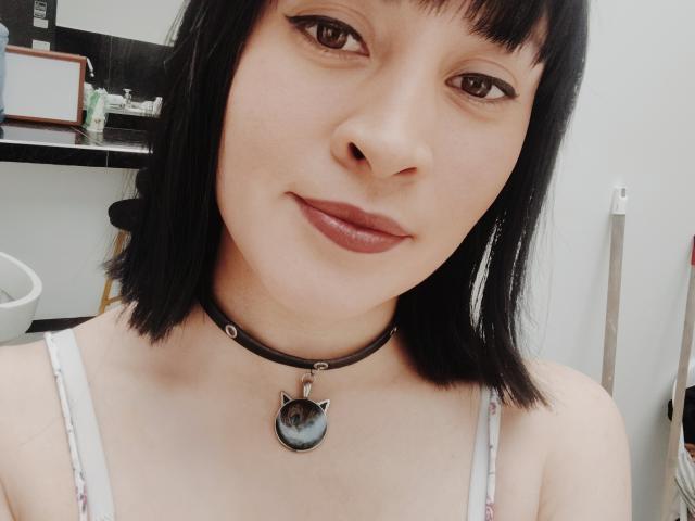 IssisDiosa tiene la piel clara, un corte de pelo bob negro, ojos marrones, y lleva un collar negro con un dije en forma de ojo de gato, sonriendo con lápiz labial rojo, y está vestida con una camiseta blanca sin mangas..