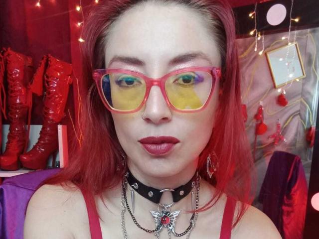 QueenScarletSecret com cabelo ruivo, óculos rosa com reflexos amarelo e azul, batom vermelho, colar preto com pingente de estrela e botas vermelhas ao fundo..