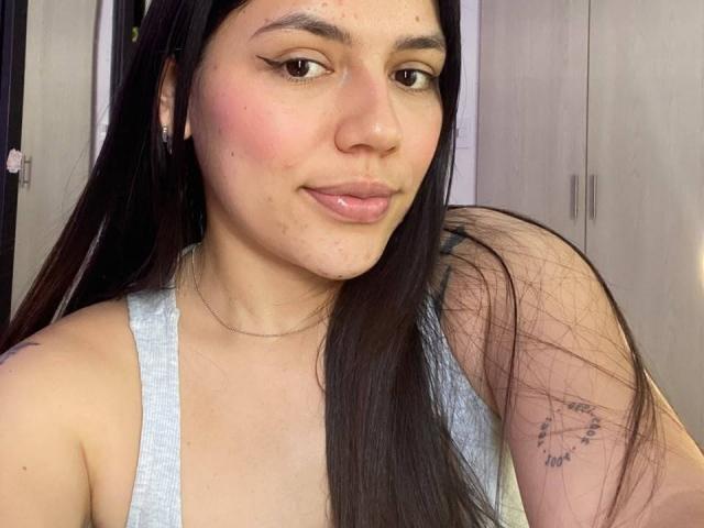 SophiaMarx with long dark hair, light skin, wearing a gray tank top, showing a tattoo on her right shoulder, smiling slightly, pink cheeks, subtle eyeliner, and small hoop earrings.