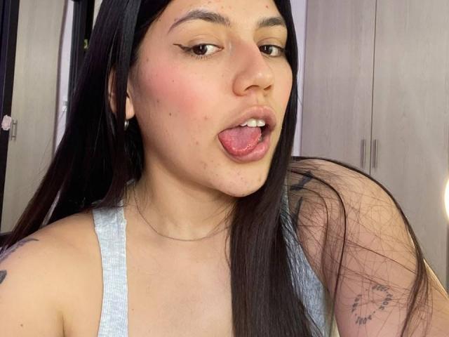 SophiaMarx has long black hair, light skin, and is sticking out her tongue with pink lipstick. She's wearing a grey tank top, showing a tattoo on her right arm.