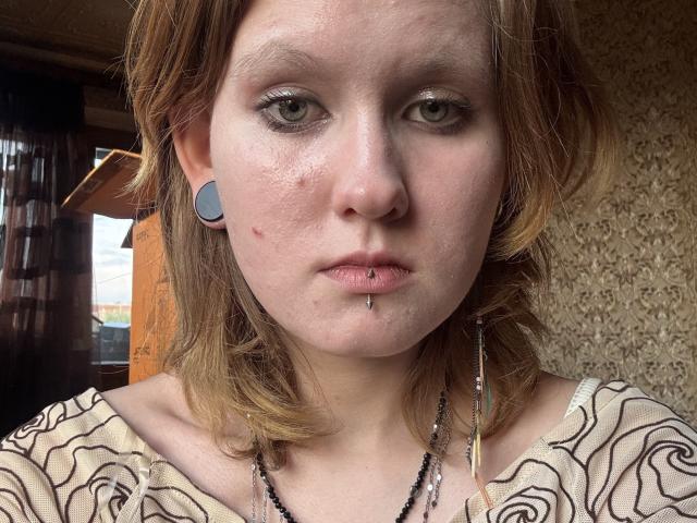 MozellContos hat eine blasse Haut, grüne Augen, ein Nasenpiercing, ein Lippenpiercing und ein Piercing an der Unterlippe. Sie trägt ein gestreiftes Hemd und blaue Ohrstecker..