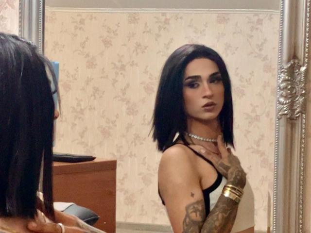 IrisIrs with black bob, tattooed arm, wearing black top, pearl necklace, gold bracelet, looking at mirror, serious expression. Mirror reflection shows IrisIrs from front.
