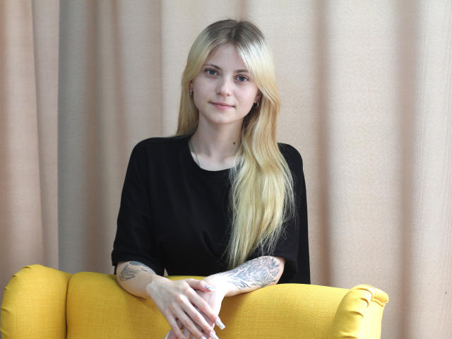 LisaOmas, con cabello rubio, camisa negra, tatuajes en el brazo derecho, sentada en un sofá amarillo, con las manos en el reposabrazos, una sonrisa sutil, piel clara, aspecto natural, iluminación suave, pose relajada..