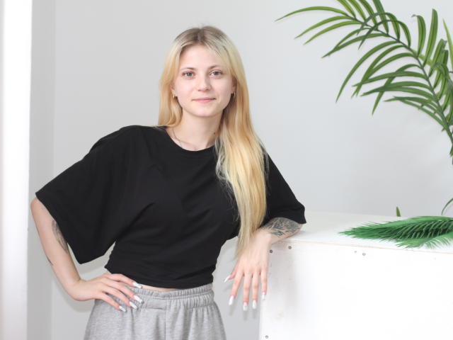 LisaOmas with long blonde hair, black tee, and grey pants, leaning on a white surface, green palm leaves in background, tattoo on right forearm, smiling, hands on hips.