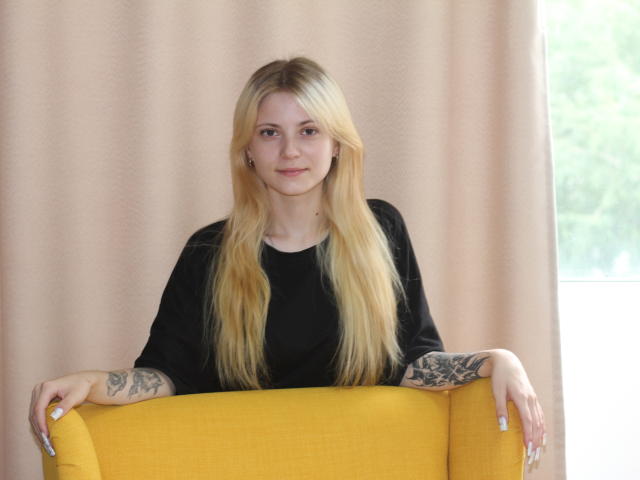 LisaOmas with long blonde hair, black top, tattoos on arms, sitting on yellow chair, hands on top, light skin, subtle smile, white nails, window on right.