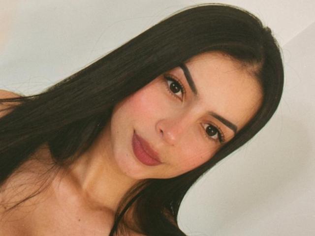 IsabellaJames with long black hair, thick eyebrows, brown eyes, and pink lipstick, smiling softly at the camera, showing her smooth, fair-skinned face and bare shoulders.