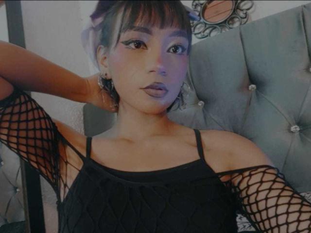 HenutmireRossi with dark brown skin, purple lipstick, and purple hair in a bun, wearing a black fishnet top, arms raised, looking at the camera.