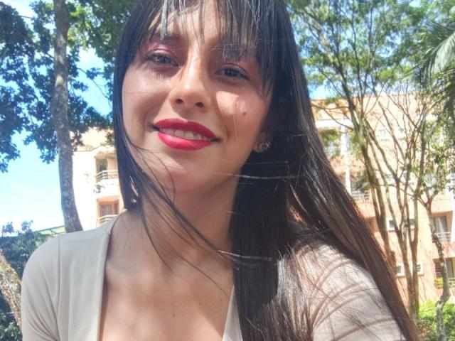 MadissonFontaine with long brown hair, red lipstick, and a smiling face, wearing a grey top, standing outside in sunlight, with trees and a building in the background.