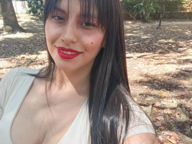 MadissonFontaine with long black hair, red lipstick, and a white V-neck top showing her tits, smiling in a sunlit forest, dirt ground, and tree shadows.