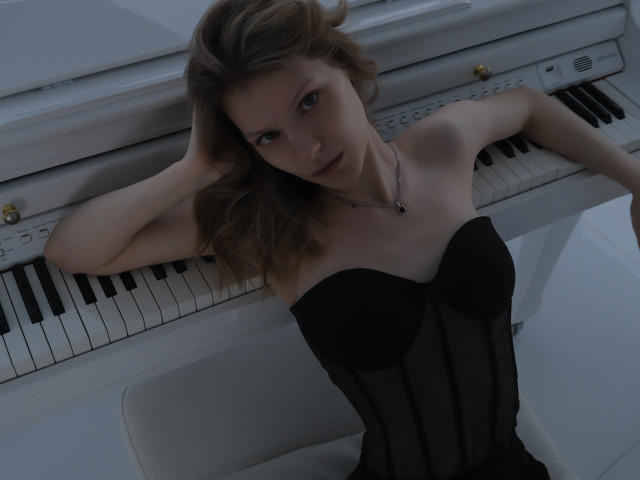 MaryHigginsx, com cabelo castanho ondulado, vestindo um corpete preto sem alças, apoiada em um piano branco, com os braços levantados, olhando para a câmera, mostrando os seios, corpo esguio, pele clara, usando um colar..