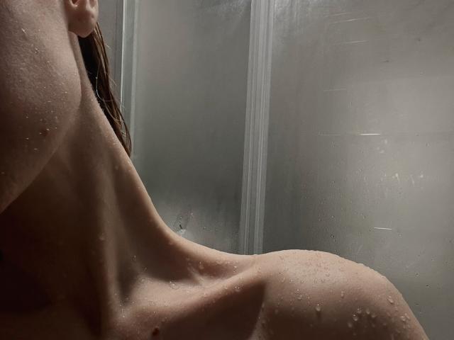 O ombro e a parte superior das costas molhados e nus de MaryHigginsx, com gotas de água, mostrando seu perfil lateral durante um banho. Seu cabelo castanho está parcialmente visível..