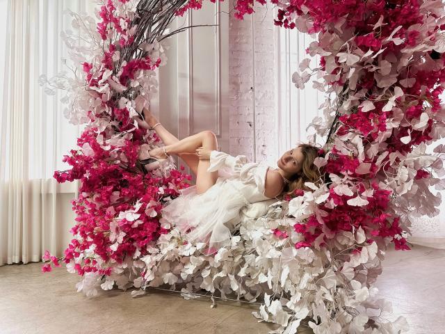 MaryHigginsx deitada em um balanço floral rosa e branco, vestindo um vestido branco, com as pernas levantadas, usando saltos altos rosa, em um quarto decorado em tons de rosa e branco..