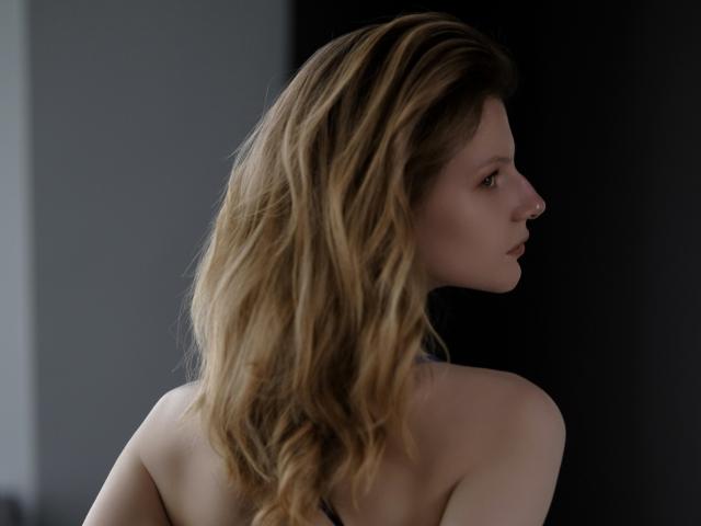 MaryHigginsx mit welligem, blondem Haar, im Profil, mit nackten Schultern, heller Haut und dezentem Make-up, blickt nach vorne mit einem ernsten Ausdruck, in einer dunklen, sanft beleuchteten Umgebung..