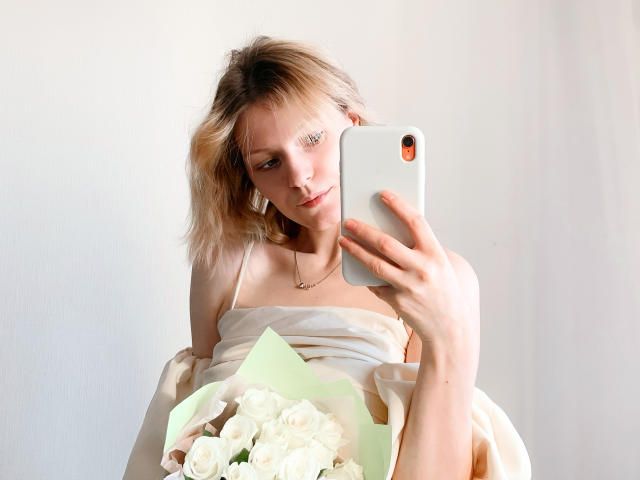 MaryHigginsx, com cabelo loiro, segurando rosas brancas em papel verde, tirando uma selfie com um iPhone branco, vestindo um vestido bege sem alças e um colar de ouro..