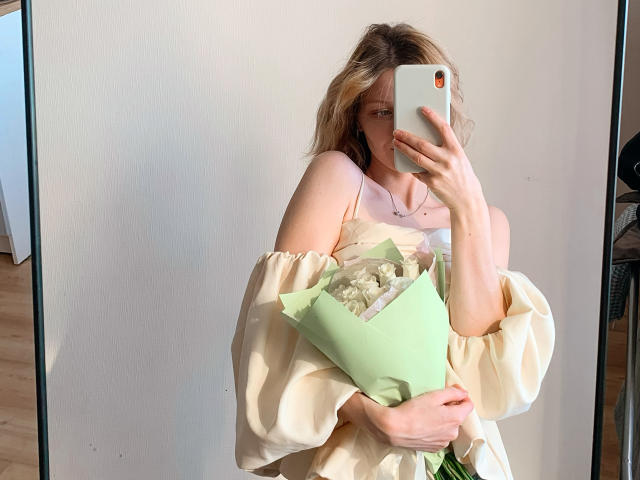 MaryHigginsx, com cabelo loiro, segurando um buquê verde, vestindo um vestido bege com um ombro descoberto, tirando uma selfie no espelho com um iPhone, pele clara, corpo esguio, seios pequenos, reflexo no espelho..