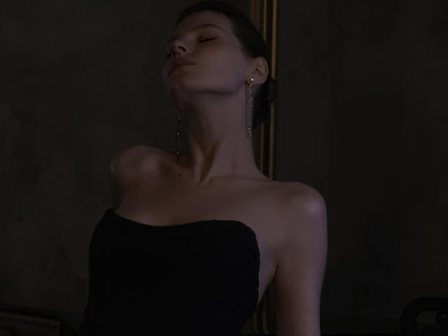 MaryHigginsx mit geschlossenen Augen, leicht geneigtem Kopf, in einem schwarzen, trägerlosen Kleid, mit silbernen Ohrringen, dunklem, zurückgebundenem Haar, bei weichem Licht, das ihre nackten Schultern und ihren Hals betont..