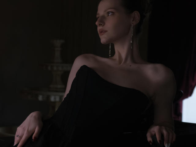 MaryHigginsx, con los hombros descubiertos, vestida con un vestido negro, con aretes, mirando hacia un lado, habitación oscura, elegante, delgada, de piel clara, maquillaje sutil, cabello oscuro, luz suave..