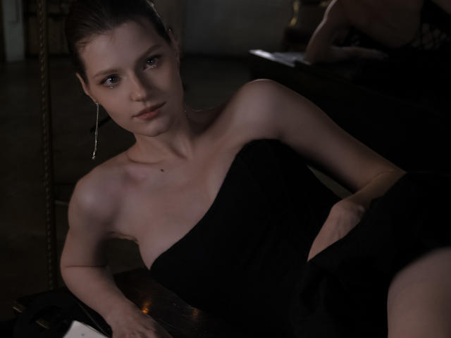 MaryHigginsx es una mujer delgada, de piel pálida, con senos pequeños, que lleva un vestido negro sin tirantes y está acostada sobre una superficie oscura, mirando a la cámara con una expresión seria..