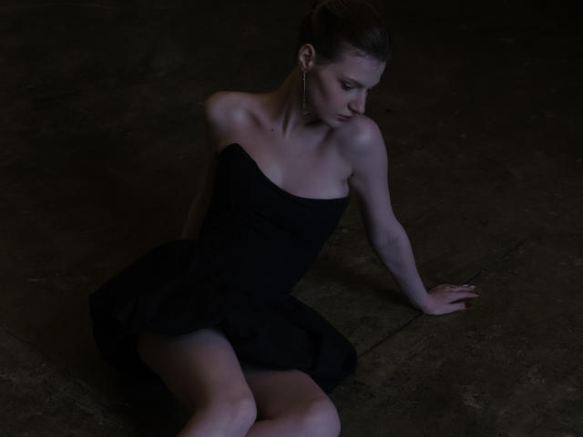 MaryHigginsx, sentada sobre el concreto, vestida con un vestido negro sin tirantes, tiene senos pequeños, piernas delgadas y está mirando hacia abajo, con un brazo detrás y el cabello castaño oscuro recogido en un moño..
