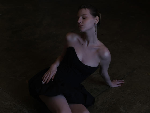 MaryHigginsx, con il seno appena coperto da un abito nero senza spalline, è seduta su un pavimento scuro, con gli occhi chiusi, un braccio all'indietro, e appare sexy ed elegante, con degli orecchini..