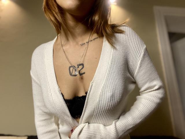 HottestDuo avec des cheveux bruns clairs, portant un cardigan blanc côtelé, laissant apparaître un soutien-gorge en dentelle noire, un collier "02", un tatouage sur la poitrine, les mains sur la taille, un éclairage subtil, un flou artistique..